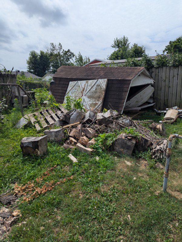 A collapsed backyard shed with its roof caved in beside a pile of logs and debris, ready to be hauled off